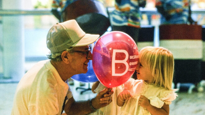 A grandfather looks through a pink clear balloon at his young granddaughter. She is a toddler with blond hair. The grandfather has a cap on and is wearing glasses. The setting is indoors.