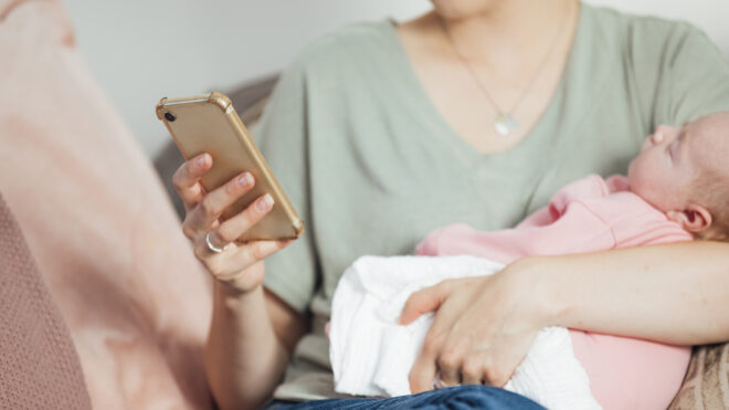 smart baby nursery, Close-up of an unrecognisable woman sitting on the sofa in her living room, holding her newborn baby in her arms while using her phone to check energy use in the home.