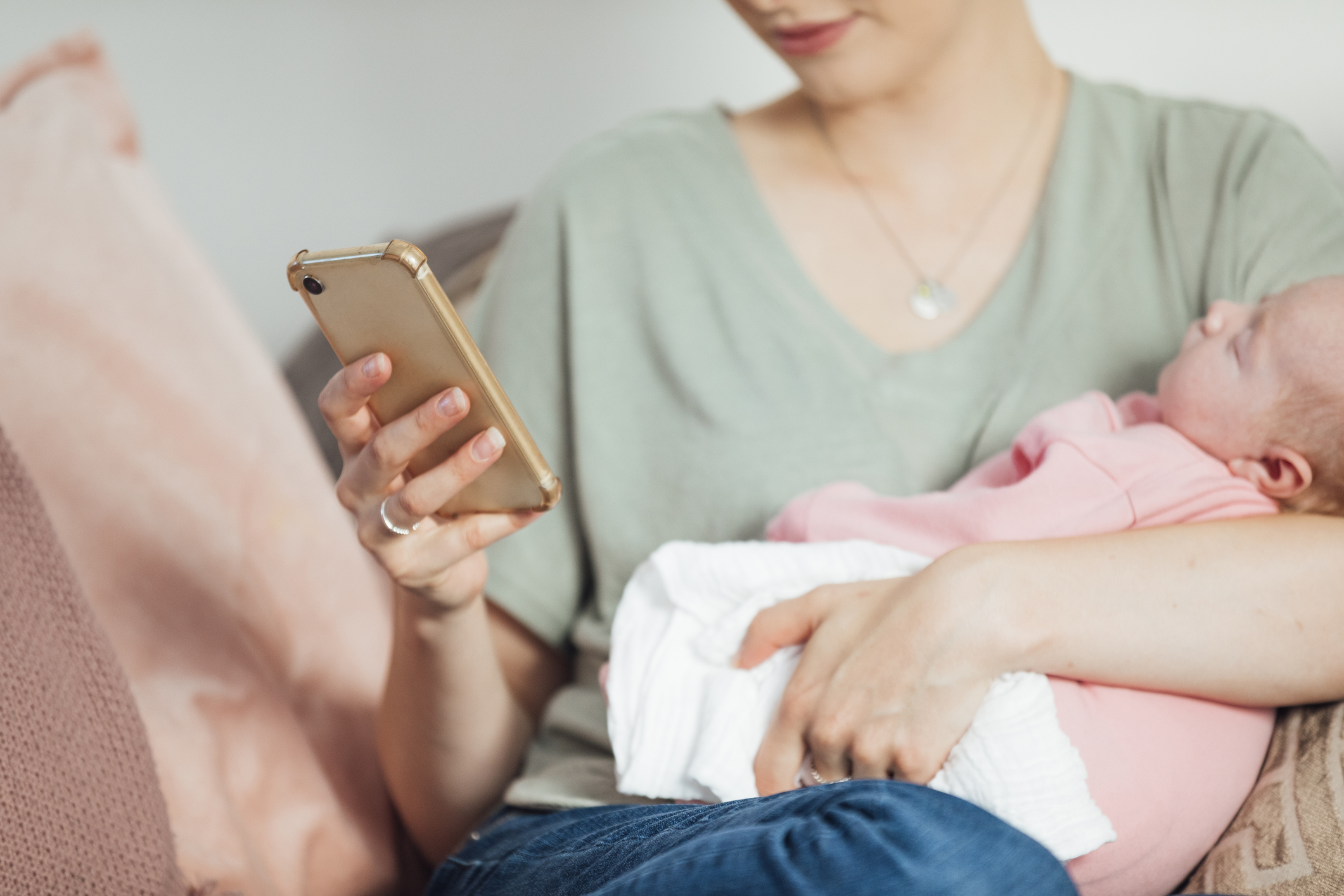 smart baby nursery, Close-up of an unrecognisable woman sitting on the sofa in her living room, holding her newborn baby in her arms while using her phone to check energy use in the home.