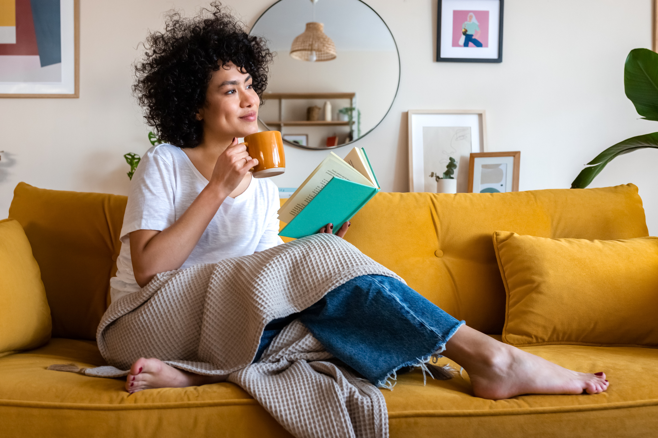 Pensive relaxed African American woman reading a book at home, drinking coffee sitting on the couch.